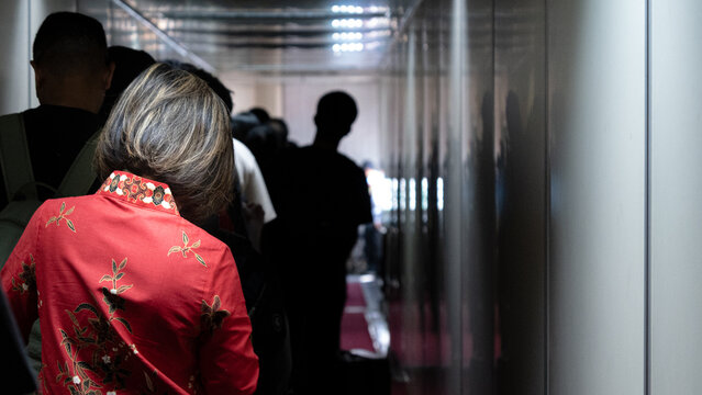 a woman wearing a traditional red batik patterned shirt stands among a queue of travelers in a narrow terminal walkway heading toward the aircraft entrance