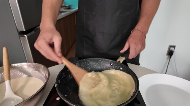 The hands of a man who roasts pancakes. Close-up. Flips a pancake with a wooden spatula on an induction hob, home cooking