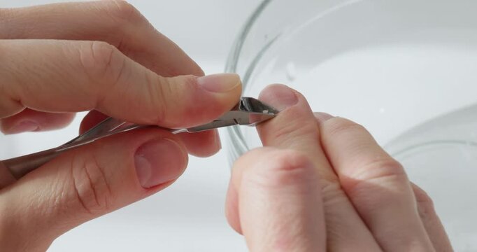 Close up of a woman's hands trimming cuticles with nippers over a bowl of water on white background