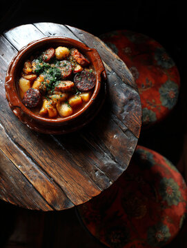 A hearty, atmospheric shot of a rustic potato stew with spicy chorizo sausage and herbs, served in a traditional terracotta dish on a weathered wooden table.