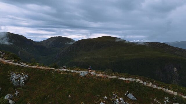 Young woman trekking uphill on paved mountain path with hiking poles and green slopes, Norwegian highlands