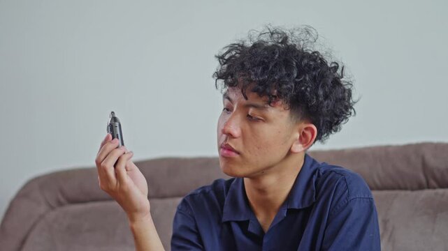 Young man with curly hair sitting on sofa and looking curiously at vape device in hand