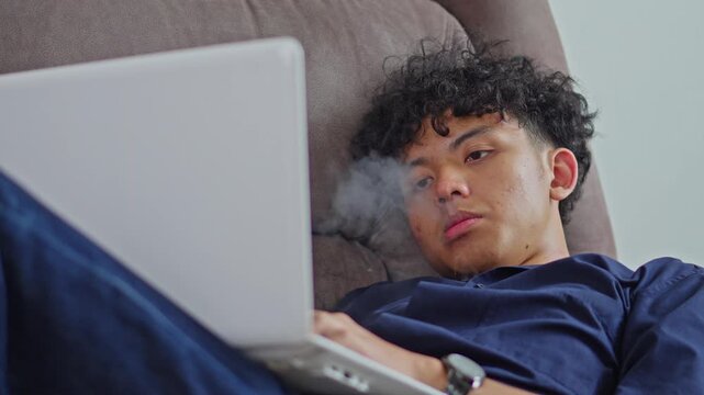 Asian male freelancer working on white laptop while vaping on couch