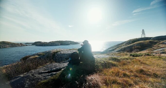 Silhouetted solo hiker sits peacefully on rugged coastal rocks with backpack beside them enjoying warm evening sunlight over Norwegian fjord. Lighthouse visible on distant hillside as golden lens flar