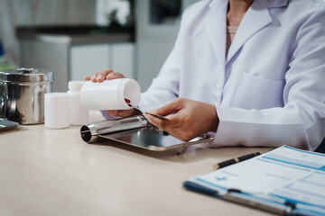 Pharmacist or lab technician in white coat counts red tablets from pill bottle onto a tray at a laboratory desk with documents nearby.