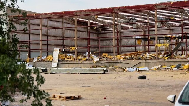 Wide view of a damaged industrial warehouse skeleton with torn insulation, metal framing and scattered debris across an outdoor construction yard in daylight.