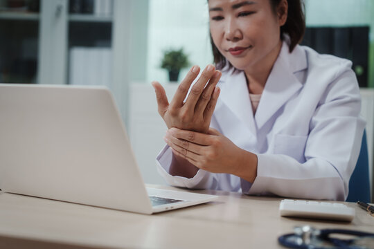 Female doctor in white coat sitting at desk, holding wrist in discomfort while looking at laptop