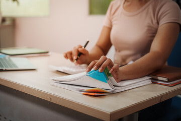 Person at desk flipping through documents with sticky tabs, calculator and laptop visible, warm indoor lighting