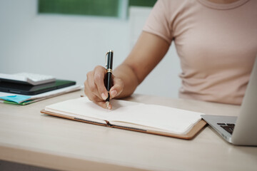 Close-up of person writing in an open notebook at a desk with laptop and documents, modern workspace, study or business planning concept.