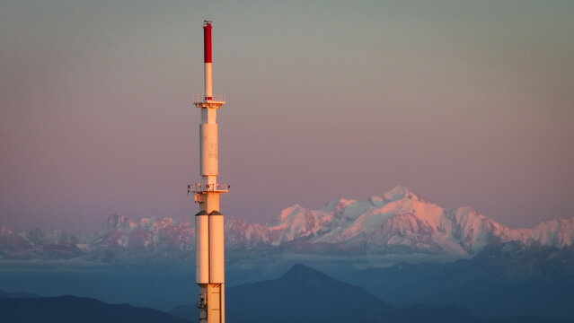 Aerial view of a tall antenna bathed in the warm glow of sunset, with snow-capped mountains in the distance, Col de la Faucille, Gex, Auvergne-Rhone-Alpes, France.