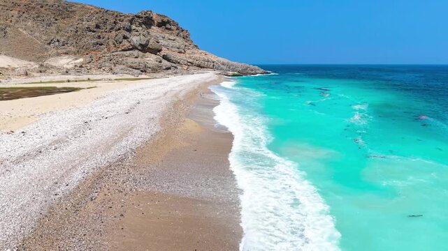 Aerial view of turquoise ocean waves crashing against a white pebble beach and rocky mountain cliffs, Saudi Arabia.