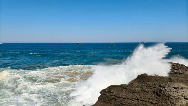 Olas del mar rompen contra formaciones rocosas en la costa, creando espuma blanca y texturas naturales en un paisaje marino din&aacute;mico y luminoso.