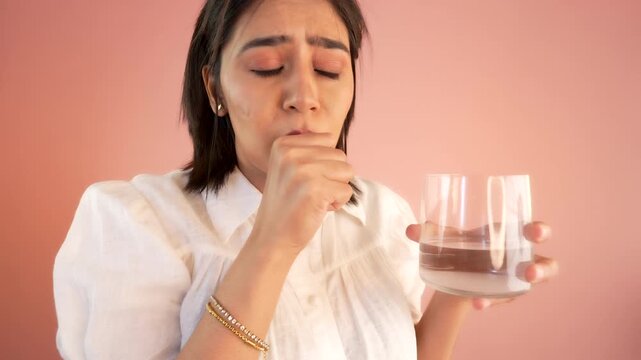 Close up of sick young woman having cough, sore throat drinking water symptoms of virus infection, isolated over light pink studio background.