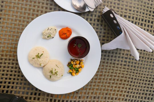 South Indian breakfast platter featuring soft steamed idlis served with traditional sambar, coconut chutney, and red chutney. The meal is accompanied by a side of boiled peas, sweet corn, and moong.