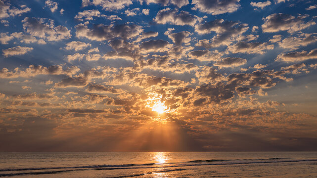 Dramatic sunrise sky with sunbeams breaking through light clouds over a sea