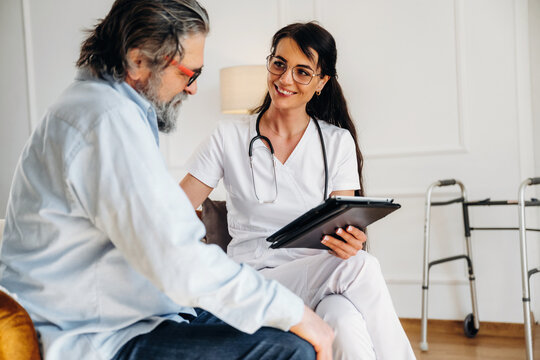Female healthcare professional in white scrubs discusses treatment options with elderly male patient seated in a well-lit room with a walker nearby
