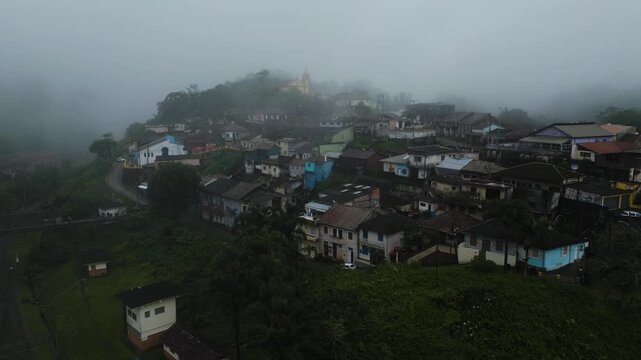 Atmospheric drone footage of Paranapiacaba historic district emerging from dense fog, showcasing colonial architecture and hillside settlement in Santo Andr&eacute;, S&atilde;o Paulo, Brazil.