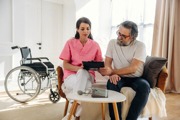 Fototapeta premium Female healthcare professional in pink scrubs discusses treatment options with senior man in casual attire, wheelchair positioned nearby in a well-lit living room setting