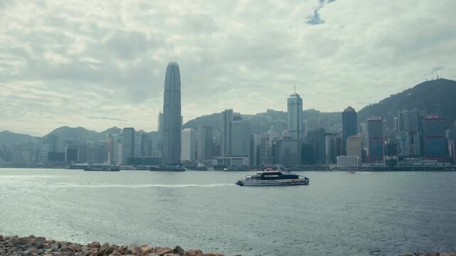 Static wide shot of a passenger ferry navigating Victoria Harbour with Hong Kong Island skyline, IFC tower, green hills and overcast sky visible from Kowloon waterfront, daytime