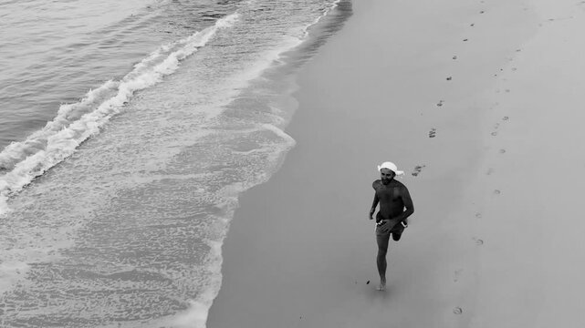Aerial drone shot of athletic man jogging along ocean shoreline on tropical beach black and white.