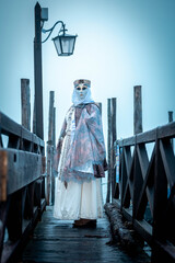 Fototapeta premium Venice, Italy - February 13, 2026: Woman in elaborate costume and mask stands on a wooden pier during Venice Carnival, lantern and fog creating a mystical atmosphere