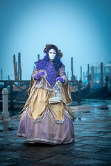 Obraz premium Venice, Italy - February 13, 2026: Woman in ornate purple and gold costume holds a lantern and fan during Venice Carnival near gondolas on a misty waterfront
