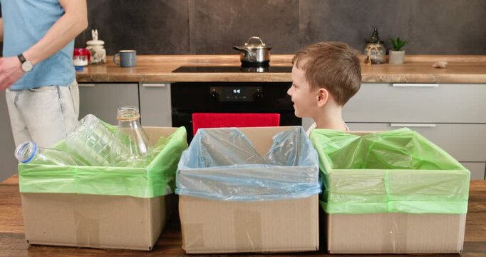 Smiling boy takes clear bottle from father holding above recycling bins in kitchen at home. Child sorts plastic bottle away from caps dropping container into proper box