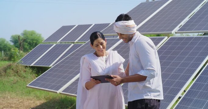Indian mature adult woman hold tab look screen visit farm field do work check record data at rural area ground place female talk speak share eco friendly solar panel power plan idea to young male