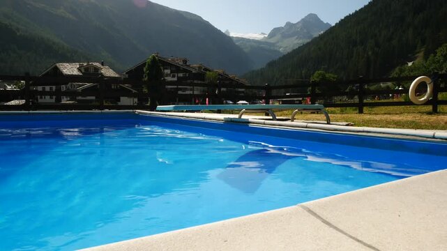 Blue calm waters of a swimming pool in a village in Aosta Valley, Italy. Mountain environment on the background, with typical buildings, mountains and blue sky.