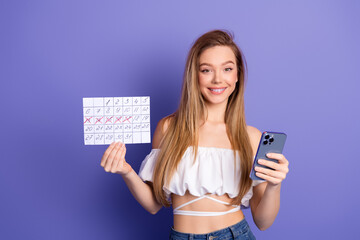 Naklejka premium Beautiful young woman holding a calendar and smartphone against a purple background, showcasing modern lifestyle and planning.