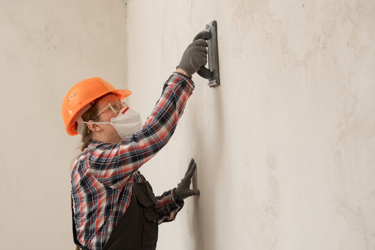 Builder woman in hardhat and protective mask sanding and smoothing walls after plastering for leveling getting it ready for painting during home renovation and construction work.