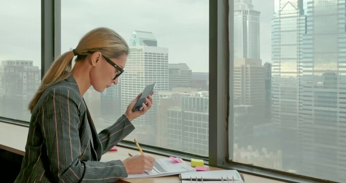Businesswoman Using Smartphone and Writing in Office with City View