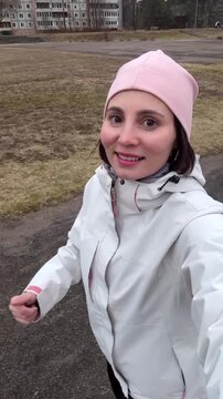 Smiling Young Woman in White Jacket and Pink Beanie Taking Wide Angle Selfie While Running on Urban Park Path &mdash; Female Jogger Filming Morning Workout on Smartphone with Overcast Sky and City Buildings