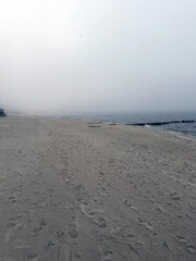 Fototapeta premium Empty sandy beach by the sea on a foggy day. Cold cloudy weather at the coast with horizon covered in mist. Nature silence and loneliness concept. Landscape scene of ocean shoreline.