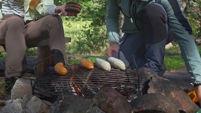 Young couple grilling sausages and burgers over campfire in sunny forest. Authentic outdoor culinary experience, sustainable living and mental wellness in a Finnish national park.