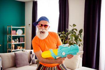 Playful grandpa in bright orange shirt aims a giant water gun indoors creating a funny cozy home...