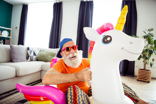 Funny grandpa in bright orange shirt and cap plays with inflatable unicorn indoors at home for a playful family moment