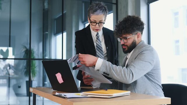 Asian woman in business attire reviews documents with African American man in gray suit at a desk in a contemporary office setting