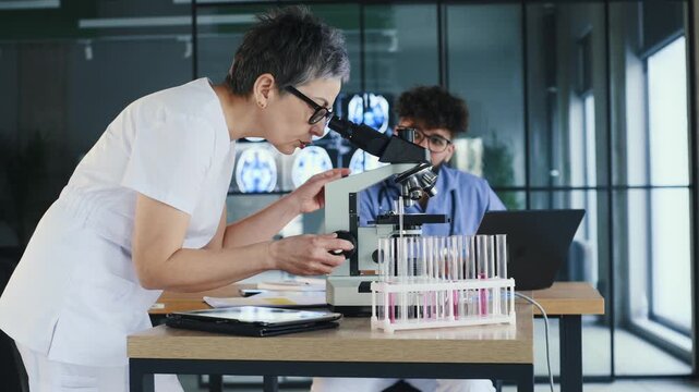 Female scientist in lab coat analyzes samples with microscope, while male coworker uses laptop in a contemporary laboratory environment