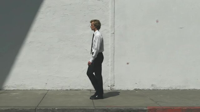 Man in business suit walking along white wall in sunlight. Businessman striding confidently through urban street. Shadow silhouette sequence showing professional commute.