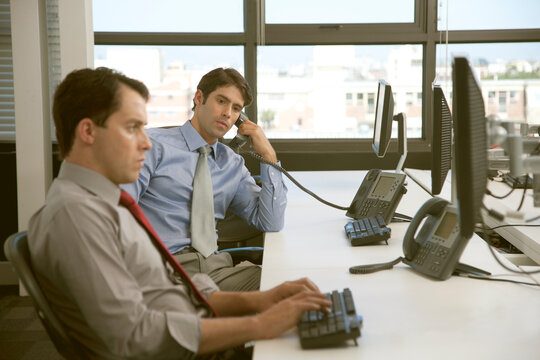 Side view of a businessman on the phone sitting next to a businessman working on a computer in an office