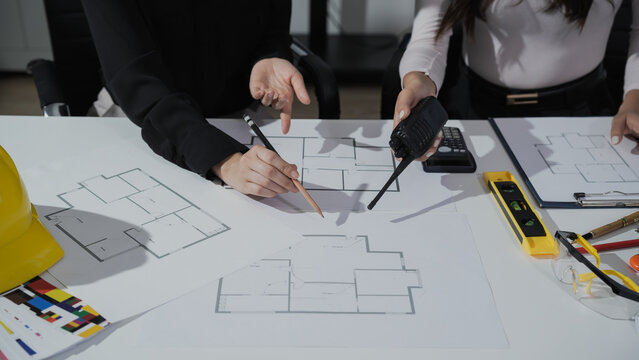 Team of architects reviewing floor plan blueprints at office desk with tools, calculator and hard hat, discussing construction design and measurements.