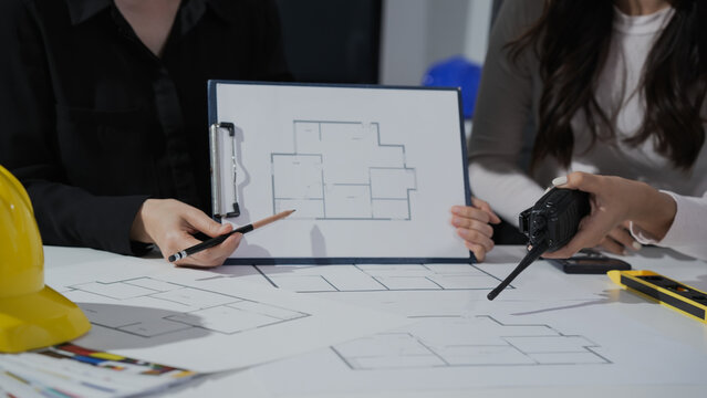 Architects review home floor plan on clipboard over desk with blueprints, hard hat and measuring tools during design meeting.