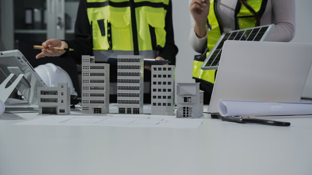 Architects and engineers reviewing building scale models with blueprints, laptop, tablet and solar panel sample in modern office, focused on construction planning and sustainable design.