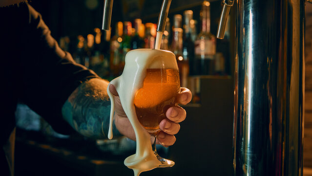 Close up of draft beer glass with thick foam spilling over bartender hand in pub. Concept of craft beer ads, beverage branding visuals, menu design asset, product promotion, bar marketing content.