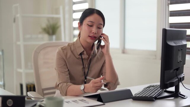 Adult Asian professional woman talking mobile phone using computer keyboard focused communication remote work office productivity business operations modern workspace