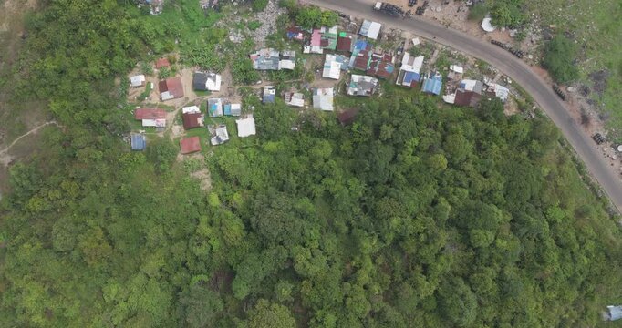 Forward aerial reveal from a hilltop of a waste sorting village featuring colorful rooftops and stacks of black bags along a winding road