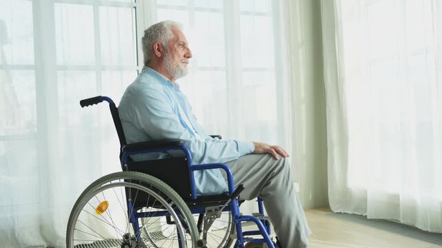 Sad senior man sitting in wheelchair looking out window in retirement home hospital. Old paralyzed man in chair for people with disability feel depressed. Lonely elderly sick man sitting on wheelchair