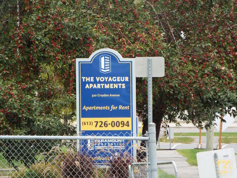Ottawa, Ontario, Canada-July 22, 2025: A blue pylon sign for "The Voyageur Apartments"on Croydon Avenue advertises available rentals in front of trees.