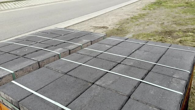 Stacked gray concrete paving stones secured with green straps on wooden pallets beside a gravel area 